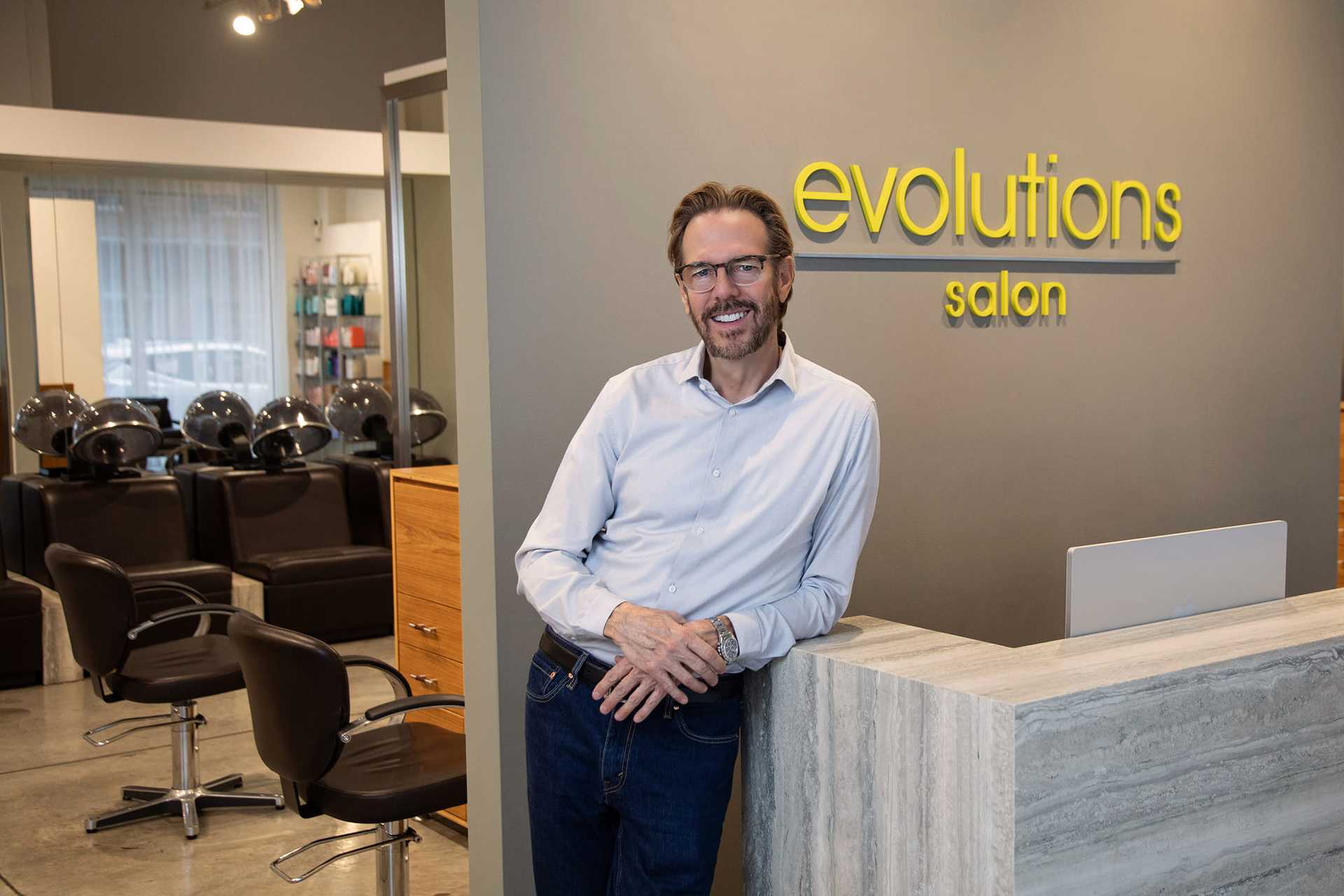 Man standing in modern salon with styling chairs and "evolutions salon" sign behind him.